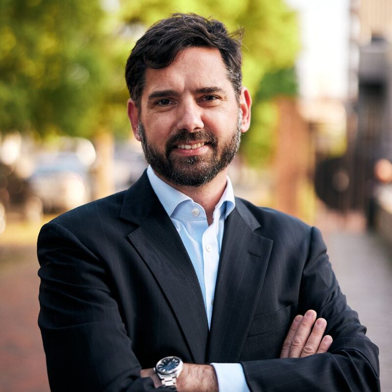 The image shows a man with a beard and short dark hair, wearing a suit jacket and a light blue shirt. He is standing outside with his arms crossed, and he is smiling at the camera. The background is blurred, but it appears to be a city street with trees.